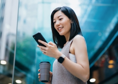 Successful young Asian businesswoman holding a reusable coffee cup, smiling after reading good news on her smartphone in contemporary office building